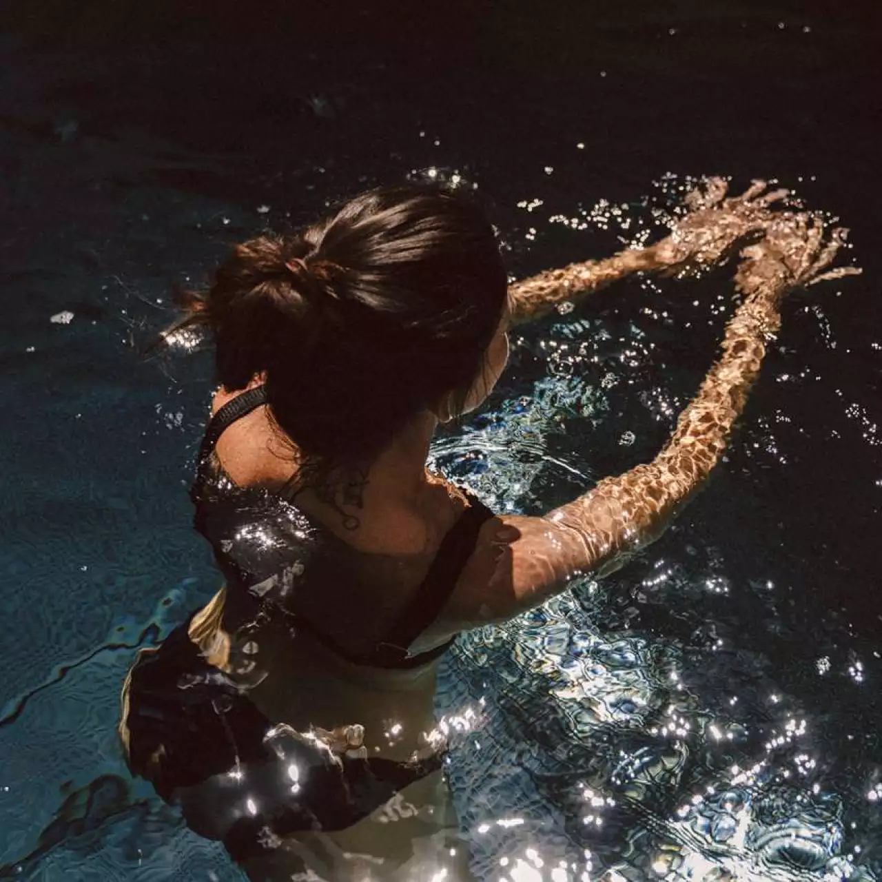 Eine Frau mit hochgestecktem Haar von hinten fotografiert, wie sie in dunklem, glitzerndem Wasser schwimmt. Das Sonnenlicht erzeugt goldene Lichtreflexe auf der Wasseroberfläche und verzerrt ihre ausgestreckten Arme unter Wasser in einem Spiel aus Licht und Schatten.